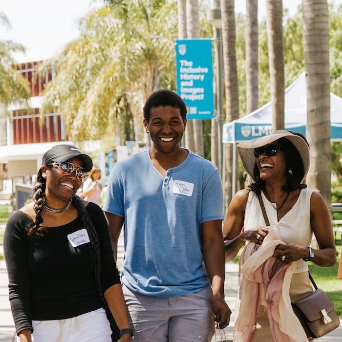 Student in a blue shirt and grey shorts laughing with his family. Adult is wearing a tan hat and dress.